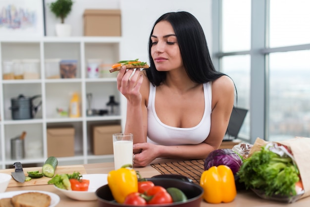 Young female standing in kitchen leaning at wooden counter, having sandwich and a glass of milk for lunch.