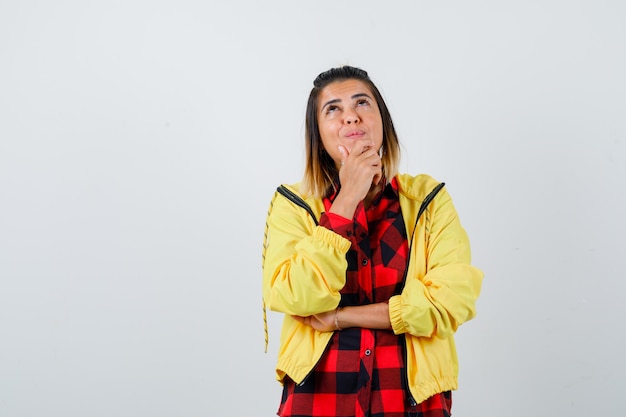 Young female standing in thinking pose while looking up in checkered shirt, jacket  and looking puzzled , front view.