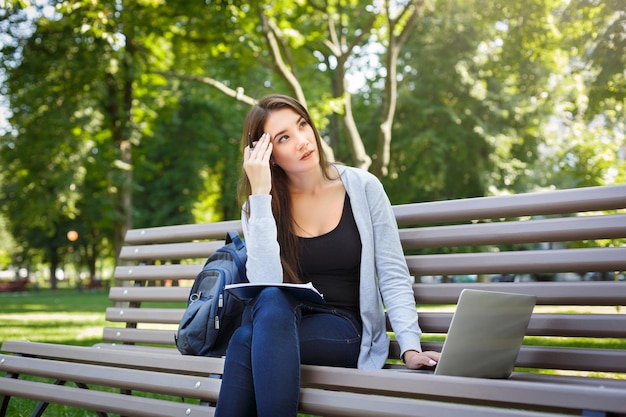 Young female student on a bench,. Thinking brunette girl doing homework in the park with laptop and textbook. Lifestyle, technology and education concept