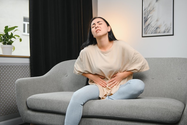 Young female suffering form stomach ache while sitting on couch at home Causes of abdominal pain include menstruation pain gastritis stomach ulcer food poisoning diarrhea or IBS Selective focus