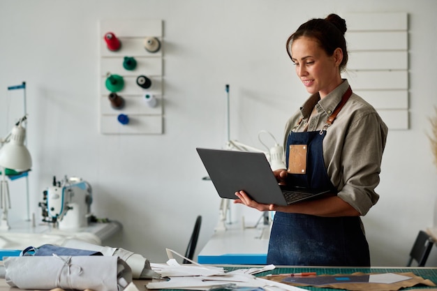 Young female tailor with laptop looking through online creative ideas