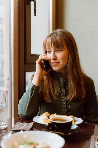 Young female talking on phone while eating dessert and drinking coffee in cafe