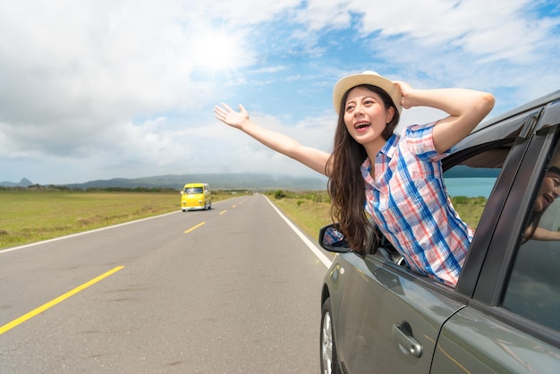 young female tourist in car on road trip showing hand happy smiling out window at summer holidays with sunlight sky and passing yellow car background.