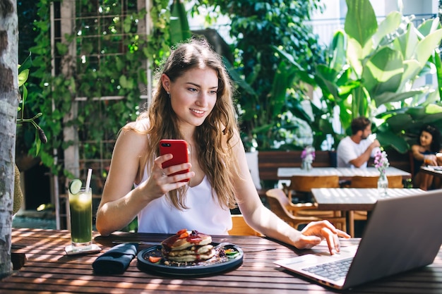 Young female using smartphone and laptop at cafe terrace