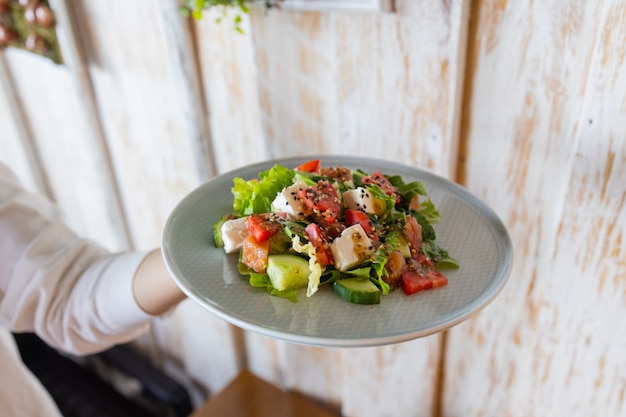 The young female waiter holding in her hands the plate with the Greek salad.