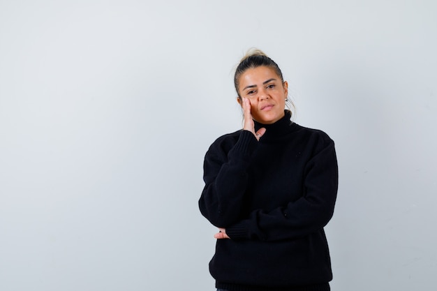 Young female with hand on cheek in turtleneck sweater and looking thoughtful. front view.