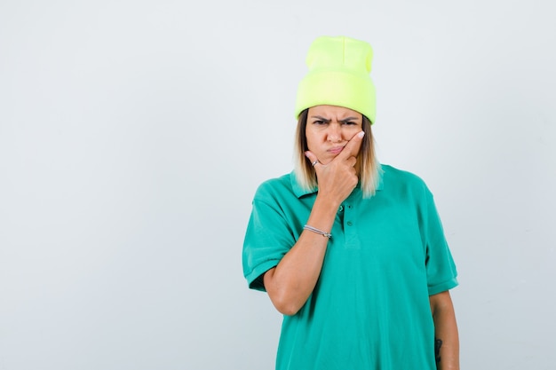 Young female with hand on face in polo t-shirt, beanie and looking bored. front view.