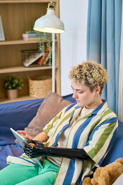 Young female with myoelectric arm using digital tablet while relaxing on sofa