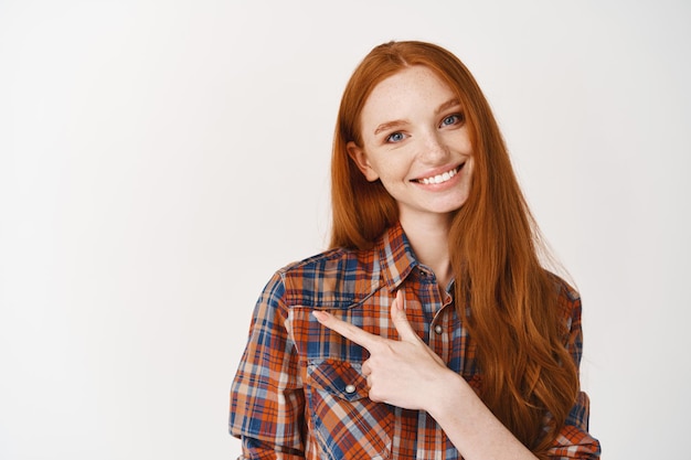 Young female with red hair and white teeth, smiling while pointing finger left, showing an advertisement on white wall