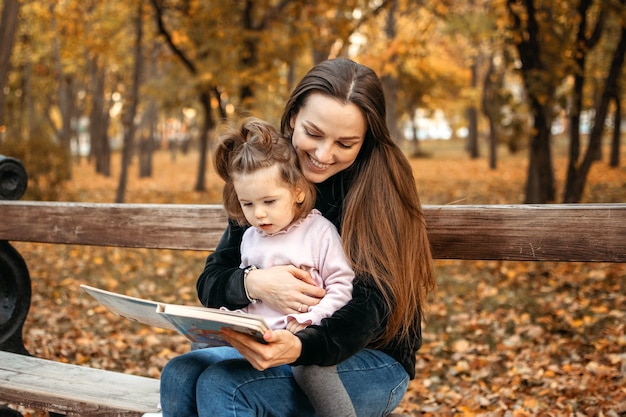 Young female woman babysitter and toddler baby girl read book in autumn park happy family mom and