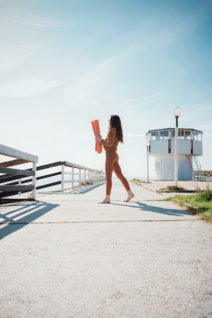 Young female yogini standing on a beach holding yoga mat in her hands