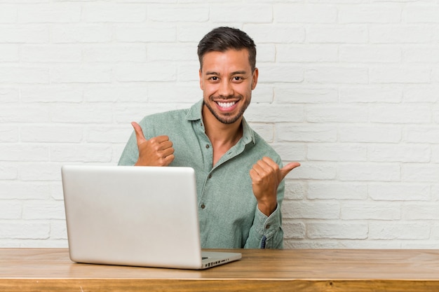 Young filipino man sitting working with his laptop raising both thumbs up, smiling and confident.