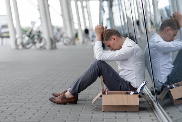 A young fired office worker is sitting under his office. Next to him is his stationery. The man is stressed.