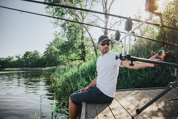 Young fisherman fishing on lake or river. Guy sit alone at water pier and look back on fish-rods. Adjust them for water hunting. Fisherman keeping legs in water.