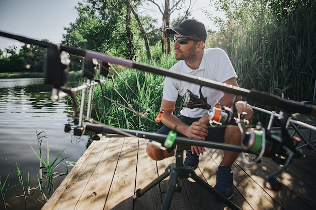 Photo young fisherman fishing on lake or river. serious concentrated professional adult guy sitting in squat position at fishing rods and looking at water. waiting for new fresh fish.