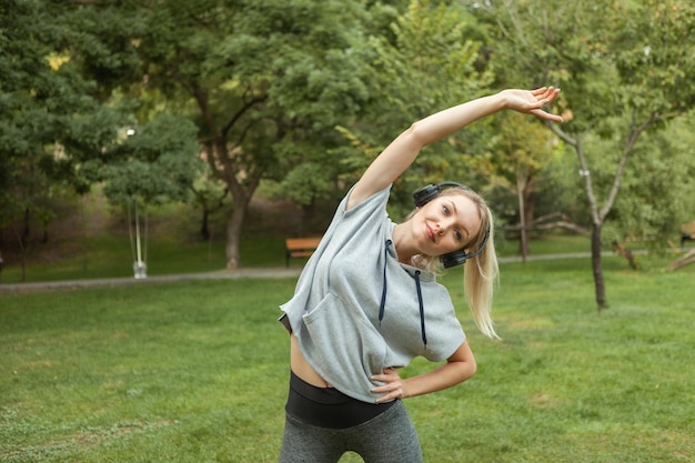 Young fit blonde woman warms up before training and listens to music on headphones in the park. Aerobica