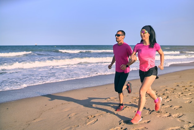 Young fit couple running on the beach during sunrise. Healthy start of the day. Wearing pink and black sportswear. Running by the sea.