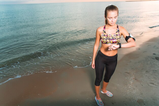 Young fit girl looking at watch during morning exercises on beach sunrise