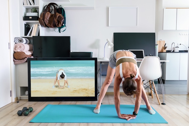 Young fit woman doing yoga stretching exercise indoor near TV screen on isolation at her home