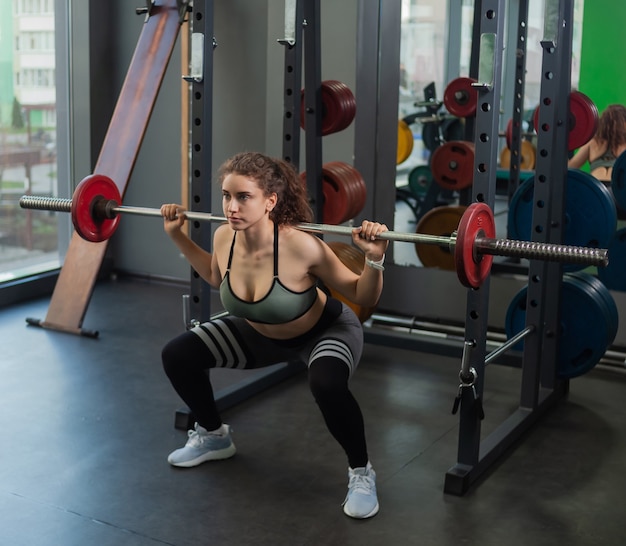 Young fit woman in sportswear crouches with a barbell on her shoulders in the gym. Healthy lifestyle concept. Hard training