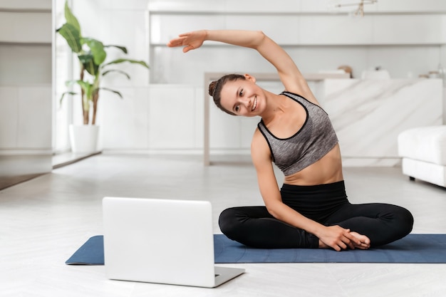 Young fit woman in sportswear practicing yoga at home with online training on laptop, watching virtual classes and tutorials