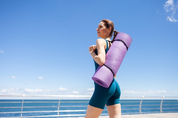 Photo young fit woman with yoga mat stands on the embankment terrace on a bright sunny day