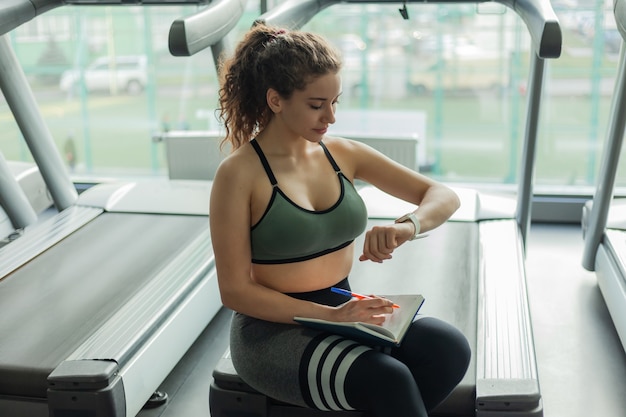 A young fit woman writes information in a training book for further progress in training in gym. Healthy lifestyle concept. Your workout plan