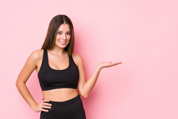 Young fitness caucasian woman isolated showing a copy space on a palm and holding another hand on waist.