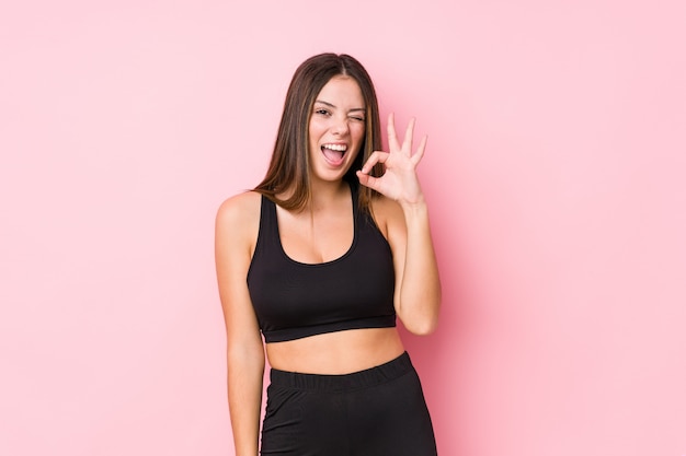 Young fitness caucasian woman isolated winks an eye and holds an okay gesture with hand.