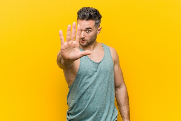 Young fitness man against a yellow standing with outstretched hand showing stop sign, preventing you.