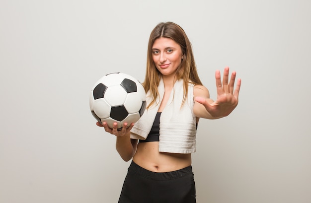 Young fitness russian woman showing number five. Holding a soccer ball.