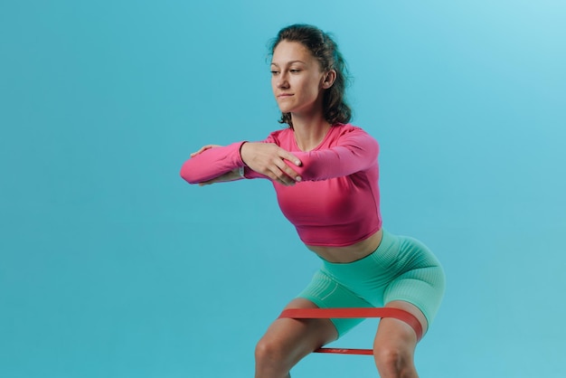 Young fitness woman doing squats with fitness loop band isolated on blue background