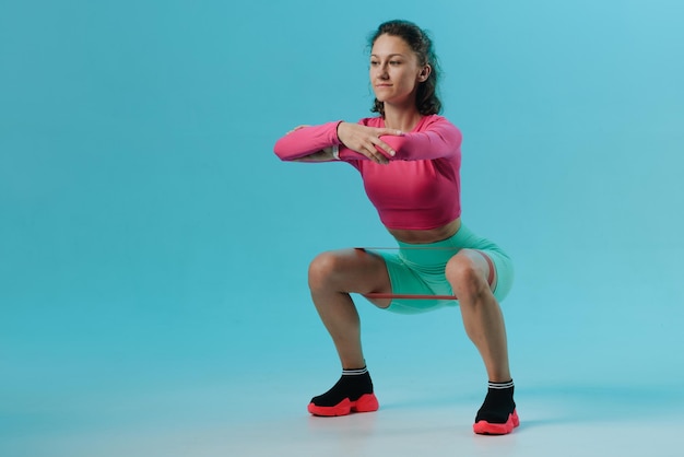 Young fitness woman doing squats with fitness loop band isolated on blue background