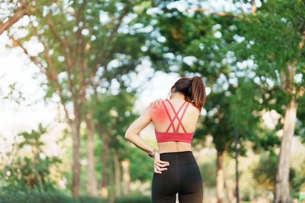 Young fitness woman holding her sports injury shoulder and neck muscle painful during training Asian runner female having body problem after exercise outside in summer