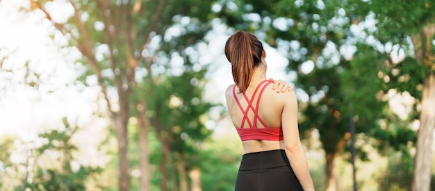 Young fitness woman holding her sports injury shoulder and neck muscle painful during training Asian runner female having body problem after exercise outside in summer