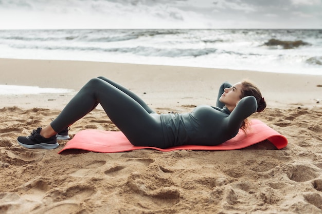 Young fitness woman trains abdominal muscles while lying on the mat on the sand at the beach