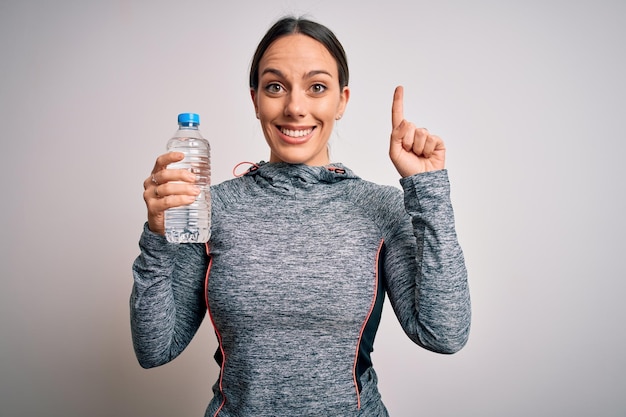 Young fitness woman wearing sport workout clothes drinking water from plastic bottle surprised with an idea or question pointing finger with happy face number one