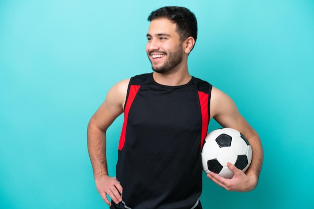 Young football player Brazilian man isolated on blue background posing with arms at hip and smiling