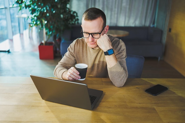 Young freelancer man in glasses sitting at a wooden table with a laptop and coffee while working on a remote project in a modern workspace