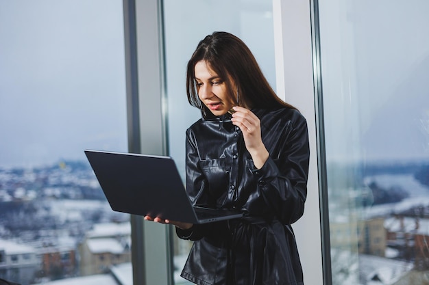 Young freelancer woman in glasses with laptop working remotely in modern workspace with large windows Remote work