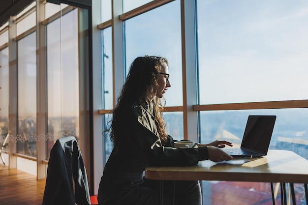 Young freelancer woman Young woman working with laptop in coworking space and talking on phone sitting in open space with cup of coffee and smiling while holding glasses