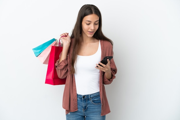 Young French girl isolated on white background holding shopping bags and writing a message with her cell phone to a friend