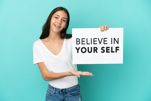 Young French woman isolated on blue background holding a placard with text Believe In Your Self and pointing it