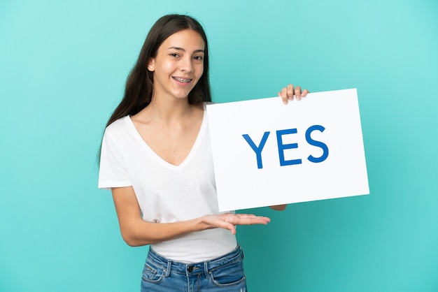 Young French woman isolated on blue background holding a placard with text YES with happy expression