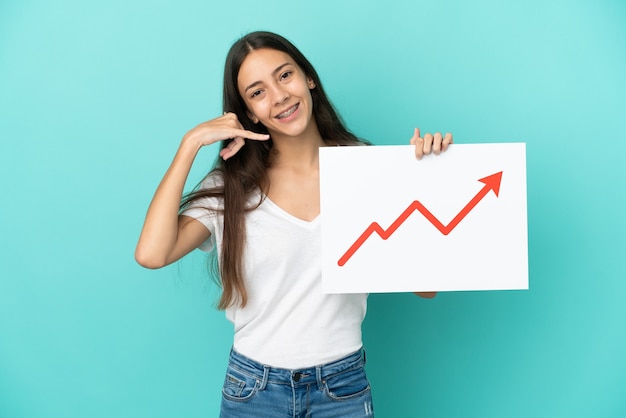 Young French woman isolated on blue background holding a sign with a growing statistics arrow symbol and doing phone gesture