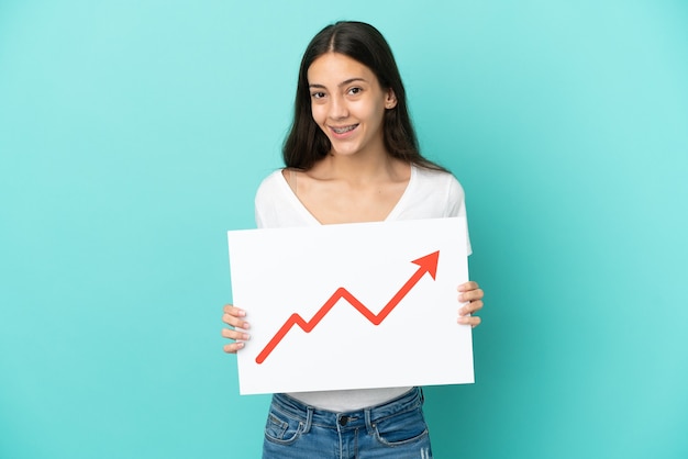 Young French woman isolated on blue background holding a sign with a growing statistics arrow symbol with happy expression