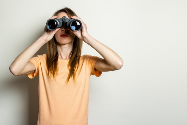 Young friendly girl in a T-shirt looks through binoculars on a light space.