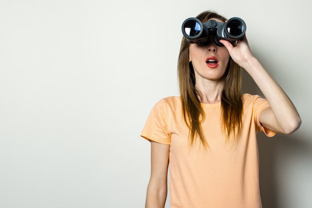 Young friendly girl in t-shirt looks through binoculars on a light space