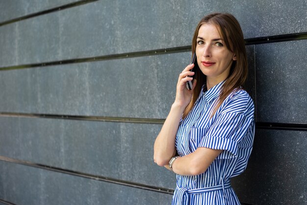 Young friendly woman in dress talking on the phone against the background of a stone wall.