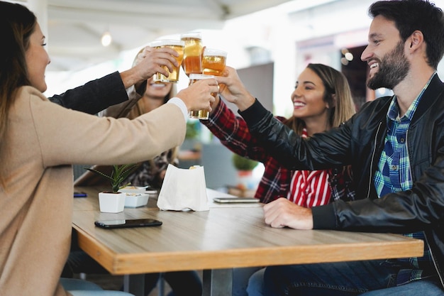 Young friends having fun cheering with beer inside bar restaurant - Focus on right girl hand